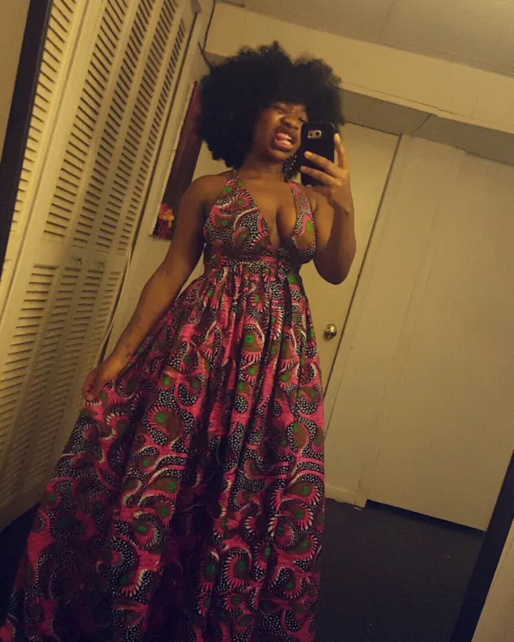 Femme noire, debout dans un couloir, portant une tenue en cuir noir avec découpes, pantalon en maille noire, chaussures à lacets, regardant vers le haut.
