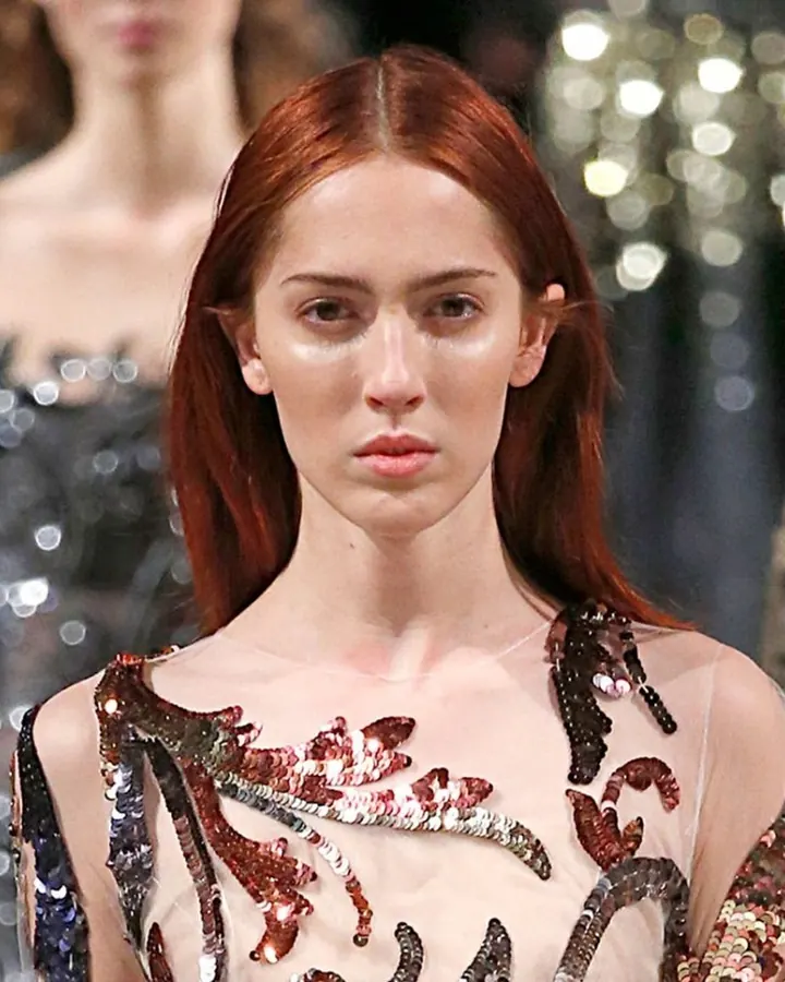 Femme aux cheveux roux, debout dans un couloir, portant une robe noire ajustée avec décolleté plongeant, cheveux blonds ondulés, tenant un sac à main.