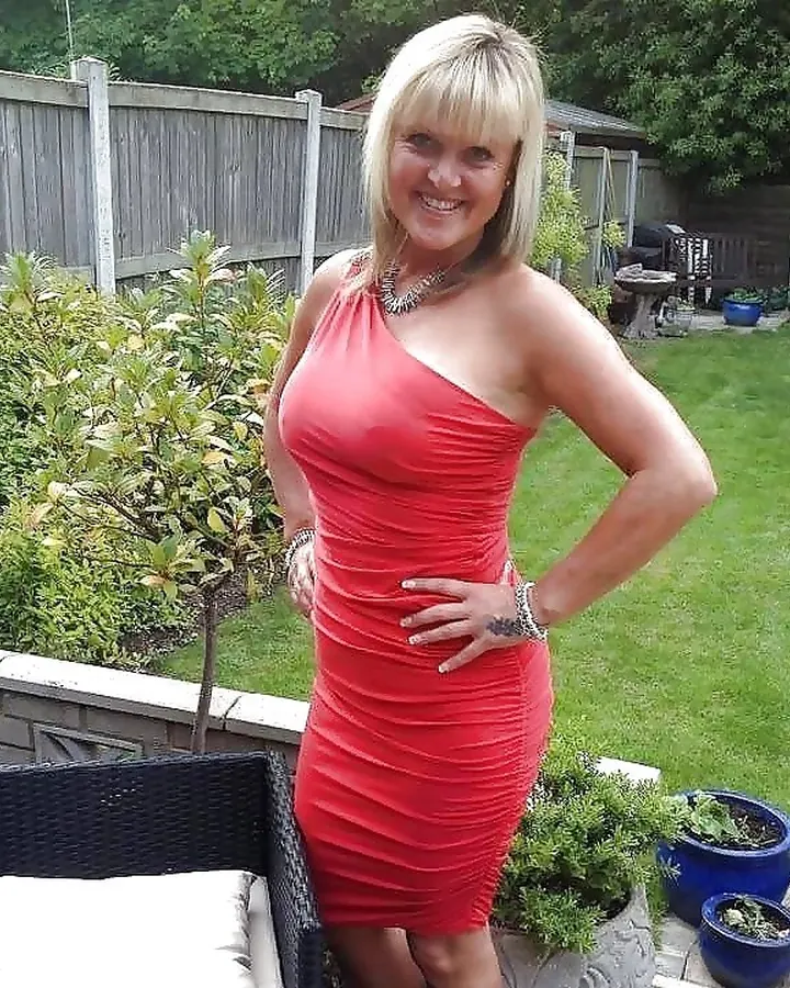 Femme blonde souriante, debout dans un jardin, portant une robe rouge ajustée, main sur la hanche, entourée de plantes et de pots bleus.