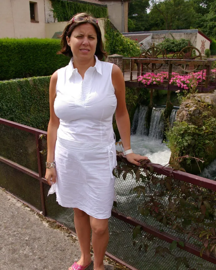 Femme aux cheveux bruns, debout sur un balcon, portant une robe multicolore, tenant un magazine, pieds nus, avec des palmiers en arrière-plan.