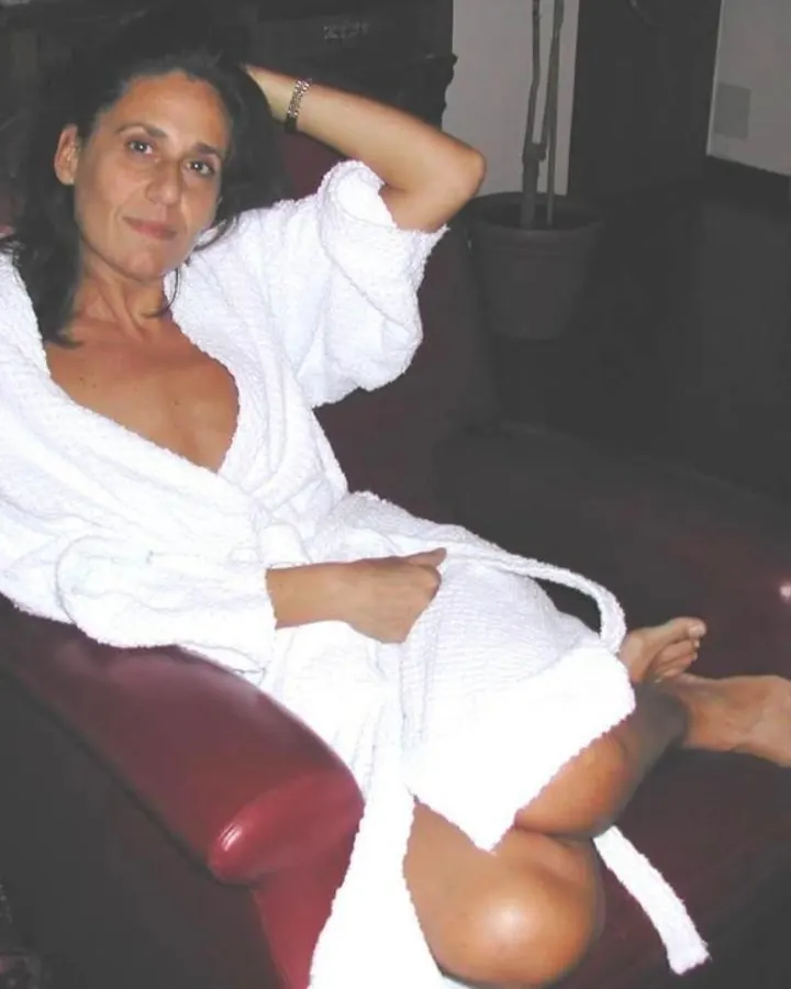 Femme assise dans un fauteuil rouge, vêtue d’un peignoir blanc, pieds nus, main posée sur sa tête.