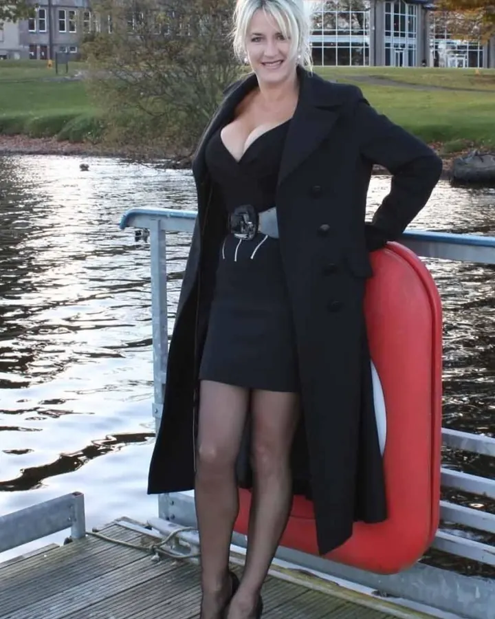 Femme blonde en manteau noir sur un ponton, près d’un lac.