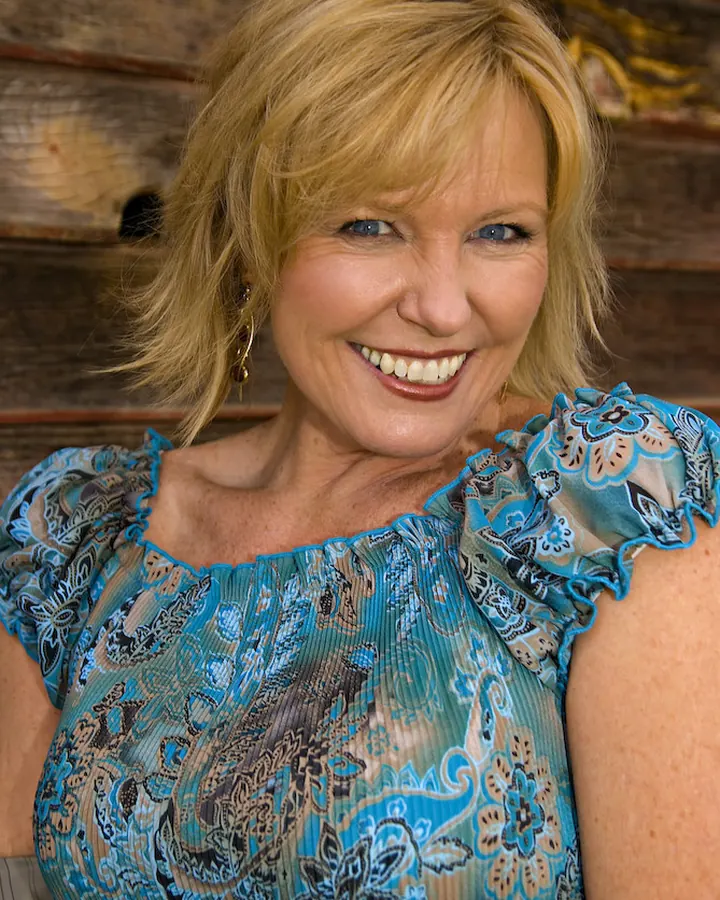 Femme aux cheveux blonds portant une robe bleue à motifs floraux, souriant devant un mur en bois.