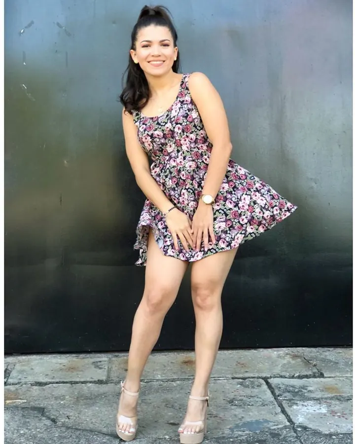 Femme en robe fleurie, souriante, posant contre un mur sombre.