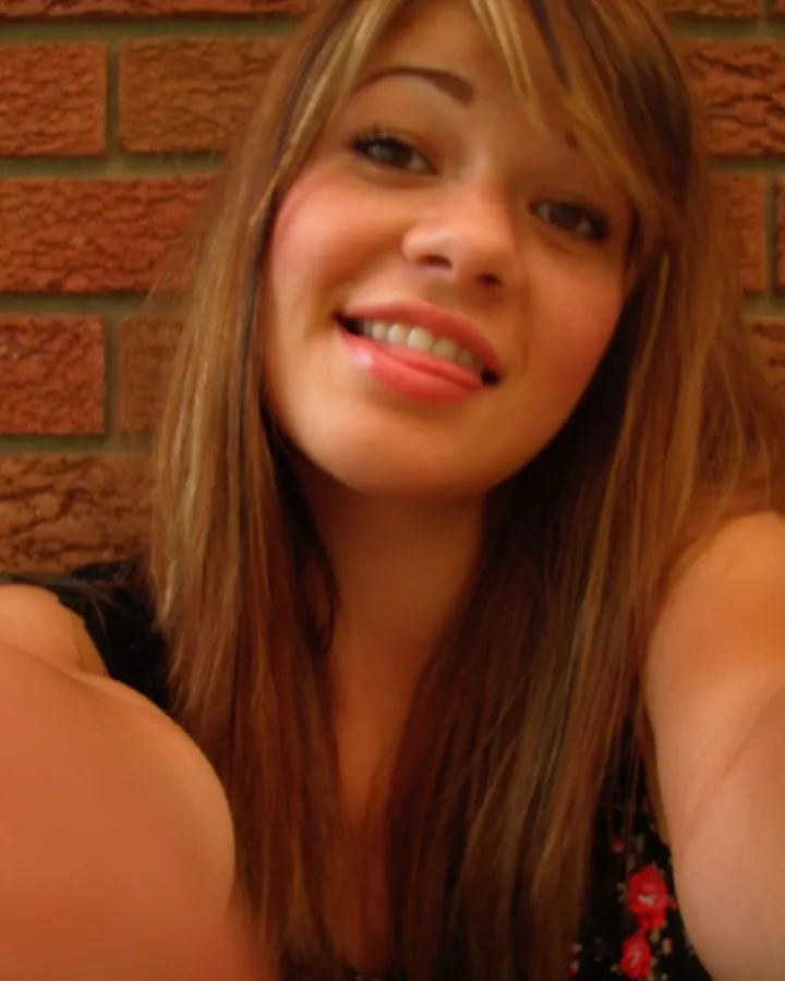 Jeune femme aux cheveux bruns, souriante, prenant un selfie devant un mur de briques.