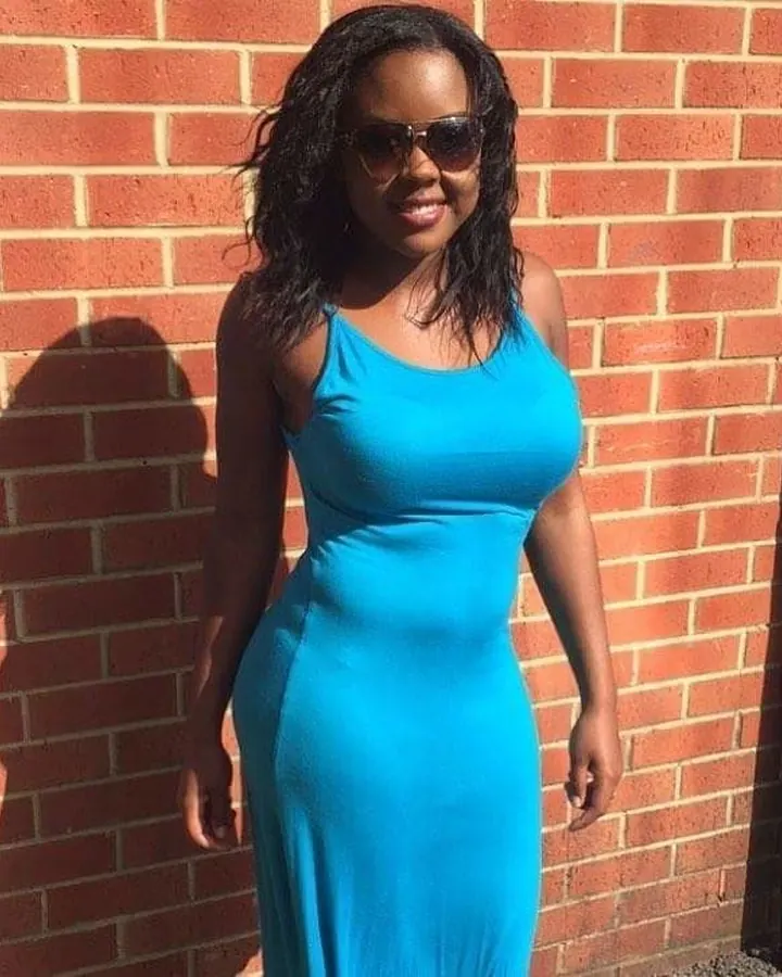 Femme noire en robe bleue, lunettes de soleil, devant un mur de briques