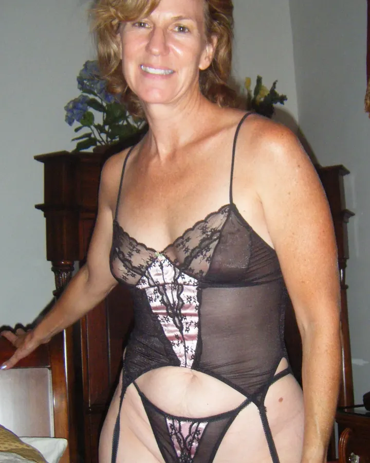 Femme aux cheveux blonds, en robe noire, debout dans un jardin, main sur la hanche, souriant.