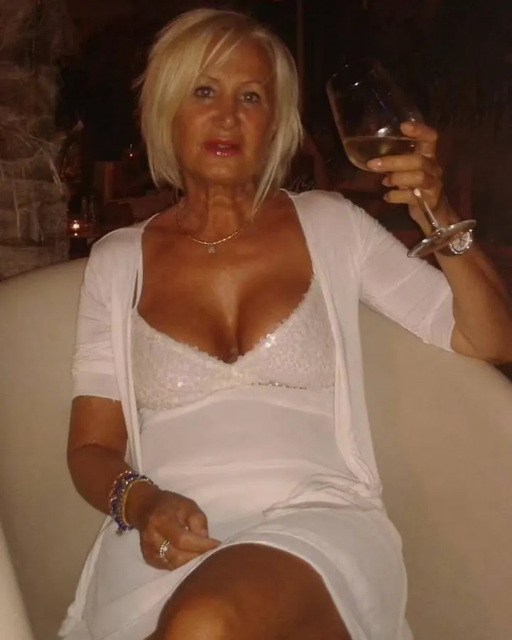 Femme blonde aux yeux marrons, en robe blanche, assise sur un canapé beige, tenant un verre de vin blanc, bijoux fins aux poignets.