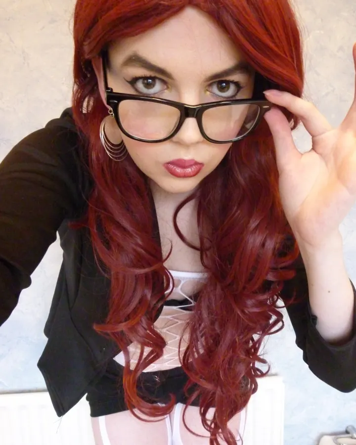 Femme rousse aux yeux marrons, en veste noire et jupe rose, collants noirs, escarpins noirs, tenant un sac à main noir, debout sur un chemin pavé dans un parc.
