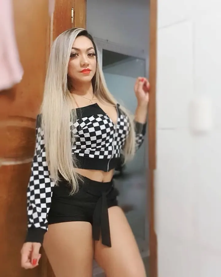Femme latino aux cheveux blonds, en robe dorée ajustée, souriante, main dans les cheveux, tenant un petit sac blanc, devant un buisson fleuri violet.