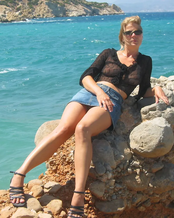 Femme blonde assise sur un mur de pierre au bord de la mer, portant un haut noir transparent et un short en jean.