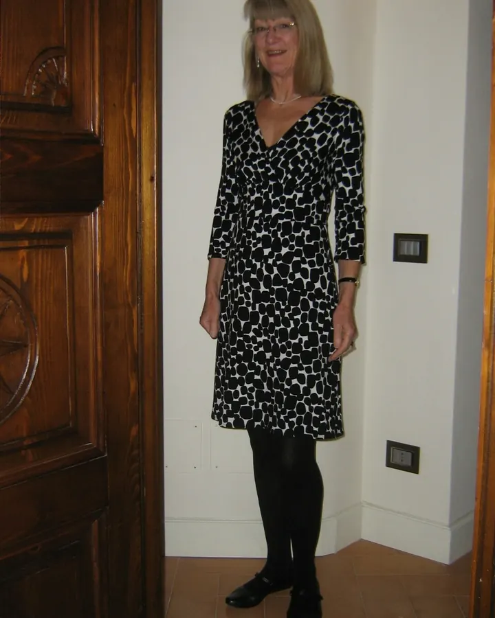 Femme aux cheveux blancs debout dans une pièce, portant une robe noire et blanche à motifs géométriques, des collants noirs et des chaussures noires.