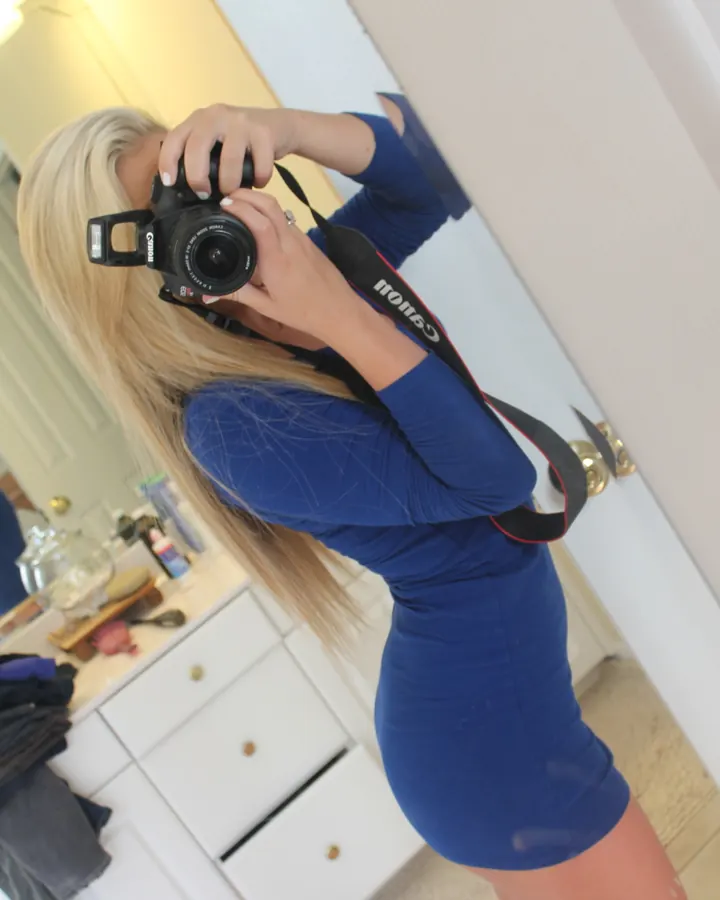Femme aux cheveux blonds prenant un selfie dans un miroir, portant une robe bleue ajustée, tenant un appareil photo Canon.