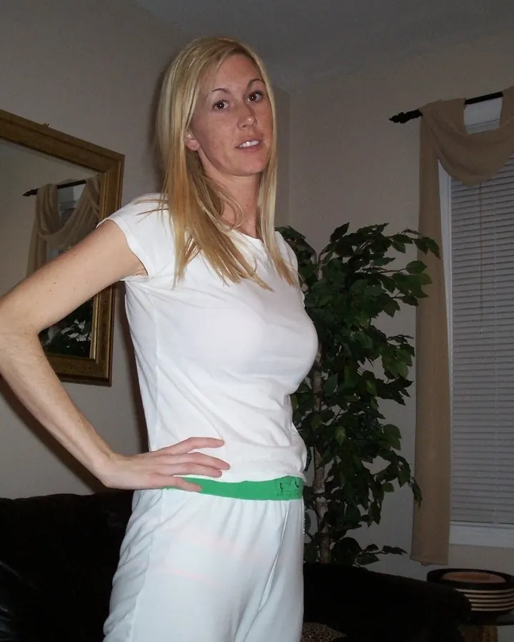 Femme aux cheveux blonds, en haut blanc, posant de face dans une salle de bain, regardant l'appareil.