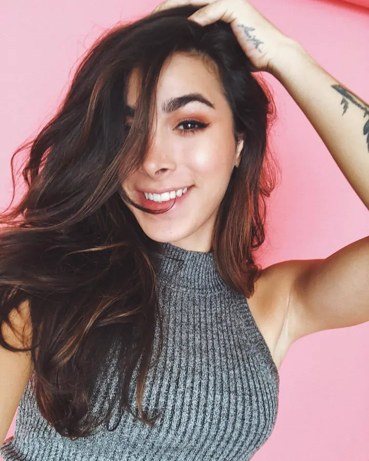 Femme aux cheveux bruns, en haut gris, posant de face dans un salon, regardant l'appareil.
