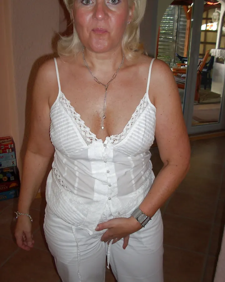 Femme aux cheveux blonds, en haut blanc et jupe en cuir, posant de profil dans une pièce, regardant l'appareil.