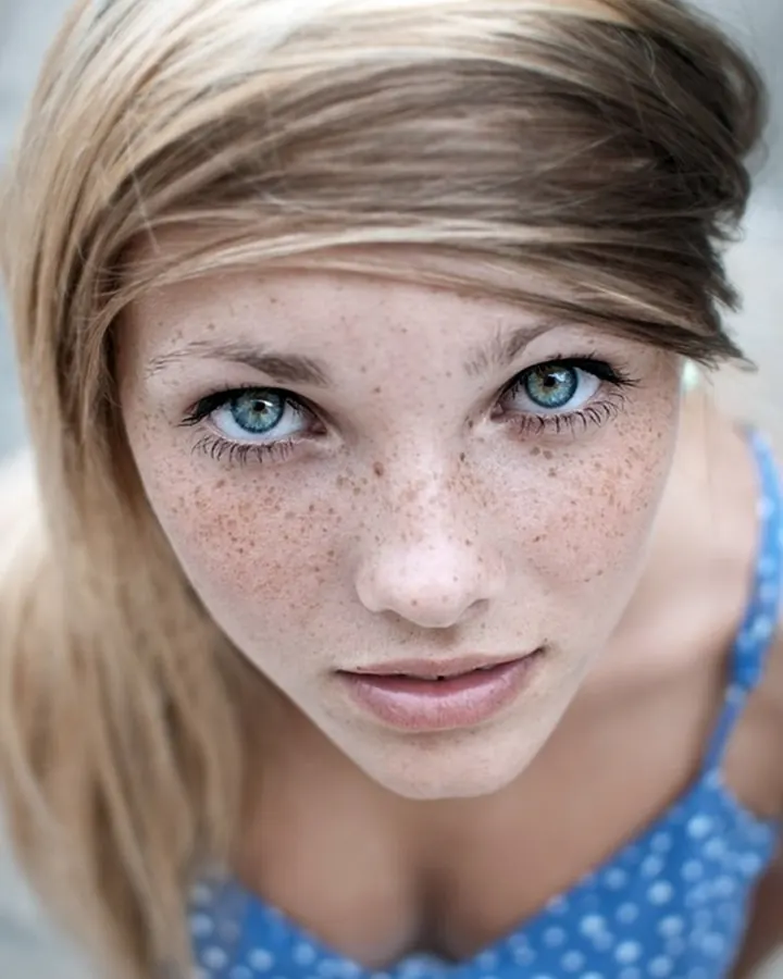 Jeune femme blonde aux yeux bleus, vue de dessus, portant un haut bleu à pois, regardant vers l'appareil.