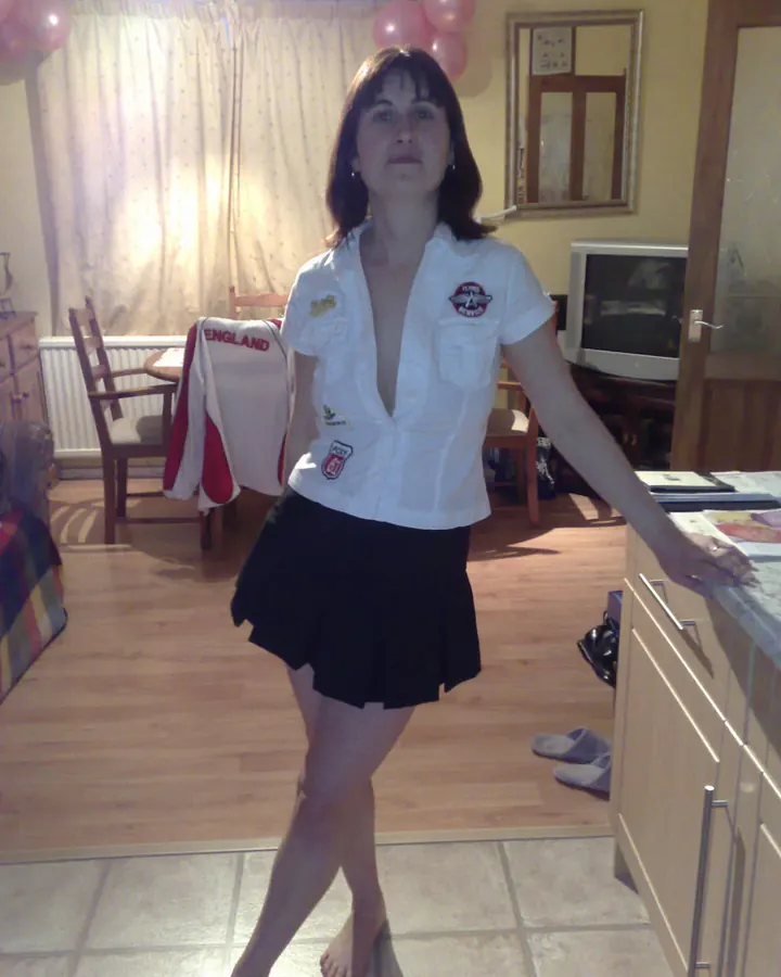 Femme debout dans une cuisine, vêtue d'une chemise blanche ouverte et d'une jupe noire courte, pieds nus, regard fixe vers l'appareil.