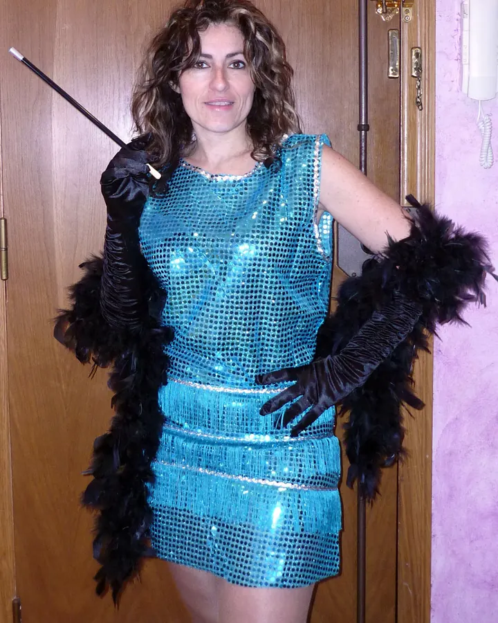 Femme vêtue d'une robe bleu turquoise pailletée, gants noirs à plumes, tenant un bâton, souriante devant une porte.