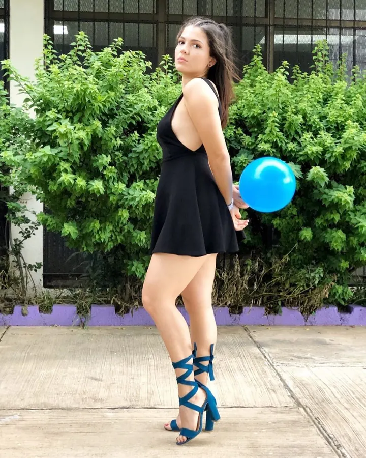Femme en robe noire courte, tenant un ballon bleu, chaussures à lacets bleues, debout devant des buissons verts.