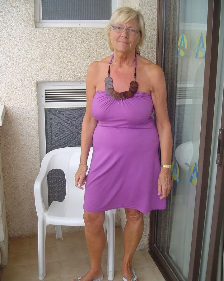 Femme blonde en robe violette, souriante, debout sur un balcon.