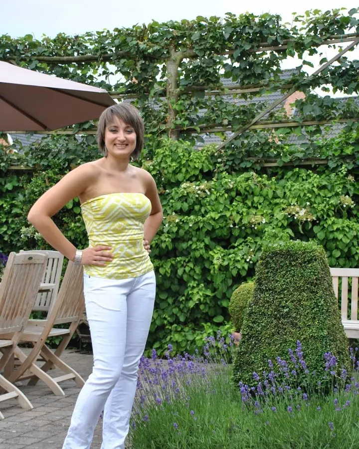 Femme en haut jaune imprimé et jeans blancs, souriante, posant dans un jardin fleuri.