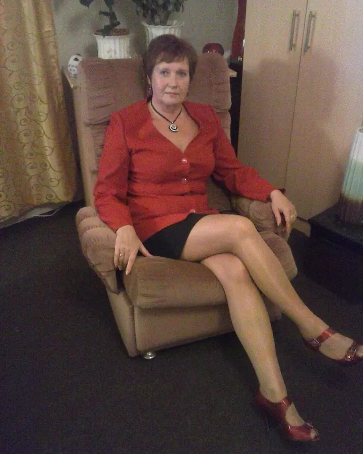 Femme assise dans un fauteuil beige, en chemisier rouge et jupe noire, les jambes croisées.