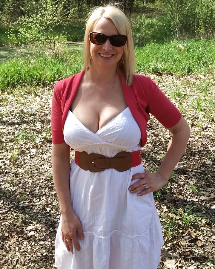 Femme blonde aux cheveux courts, en robe blanche avec cardigan rouge, souriante dans un cadre naturel.