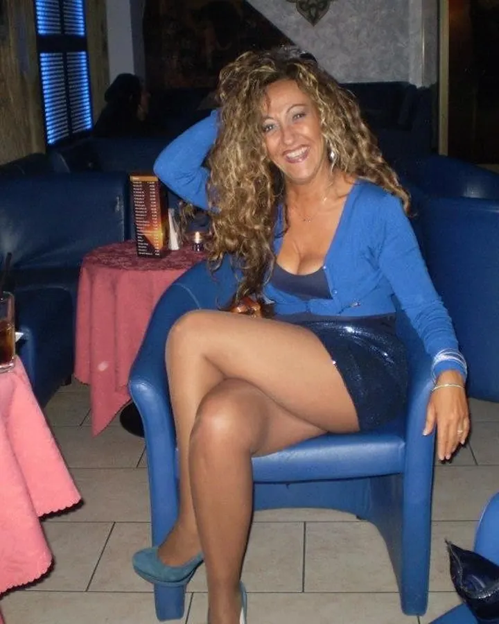 Femme aux cheveux blonds bouclés, en tenue bleue, assise sur un fauteuil dans un bar.