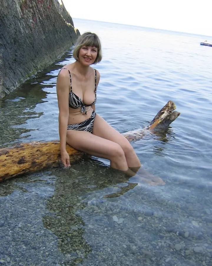 Femme aux cheveux platine en bikini noir, assise sur un tronc dans l'eau