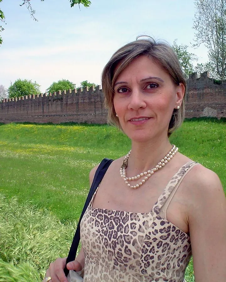 Femme aux cheveux blonds, en robe léopard, souriant devant un mur de briques anciennes avec un champ de fleurs jaunes.