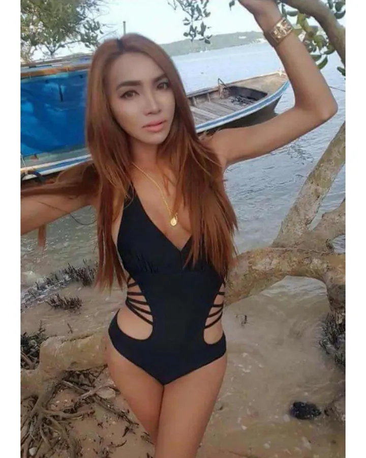 Femme asiatique aux cheveux châtain, en maillot de bain noir à découpes, posant près d’un bateau sur une plage.