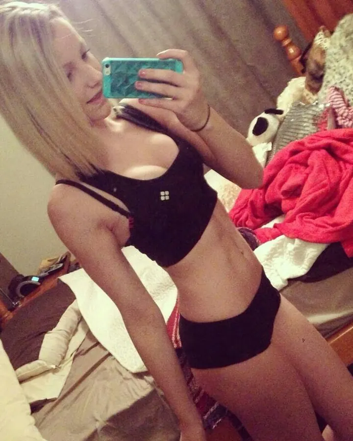 Femme blonde, en haut noir et culotte noire, prenant un selfie dans une chambre, avec un lit et des couvertures en arrière-plan.