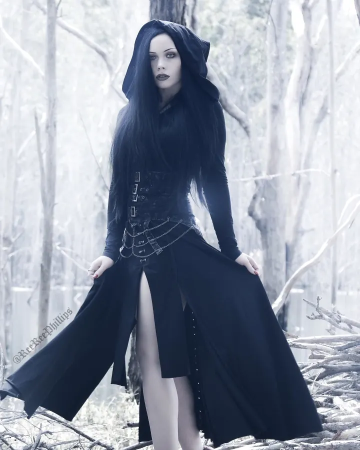 Femme aux cheveux noirs, en tenue gothique noire avec capuche, posant dans une forêt blanchie, entourée de branches mortes.