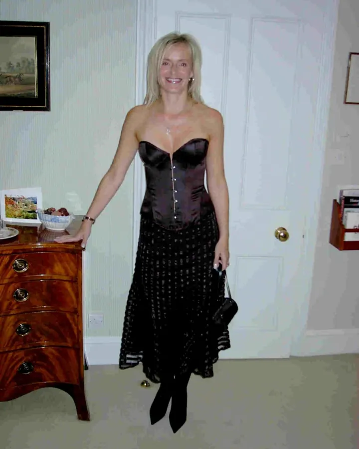 Femme blonde souriante, en robe noire bustier et jupe longue, tenant un sac, près d'une commode.