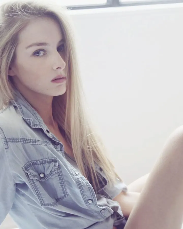 Femme blonde, assise sur un lit, portant une chemise en jean, regardant l'appareil photo.