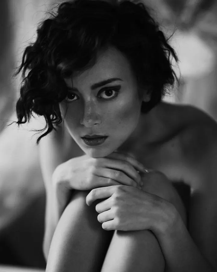 Femme aux cheveux bruns-noirs portant un haut noir, avec des boucles d'oreilles, regardant l'appareil photo avec un sourire discret.