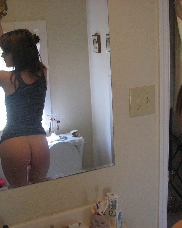 Femme aux cheveux châtains prenant un selfie dans un miroir de salle de bain, vêtue d'un haut noir et nue en bas.