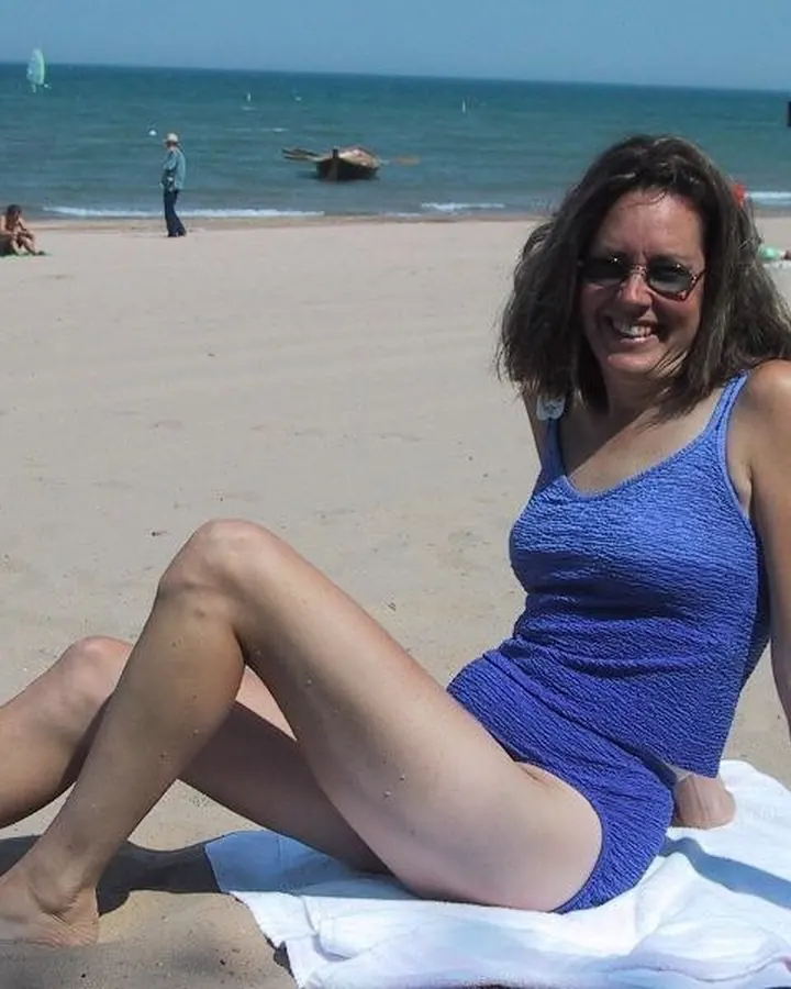 Femme aux cheveux bruns foncés, en maillot de bain bleu, assise sur une plage, souriante, avec des lunettes de soleil.