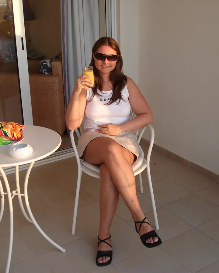 Femme mûre aux cheveux bruns portant des lunettes de soleil, assise à une table de balcon avec un verre de jus d'orange.