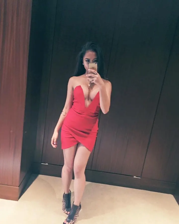 Femme arabe en robe rouge moulante, prenant un selfie devant un mur sombre.