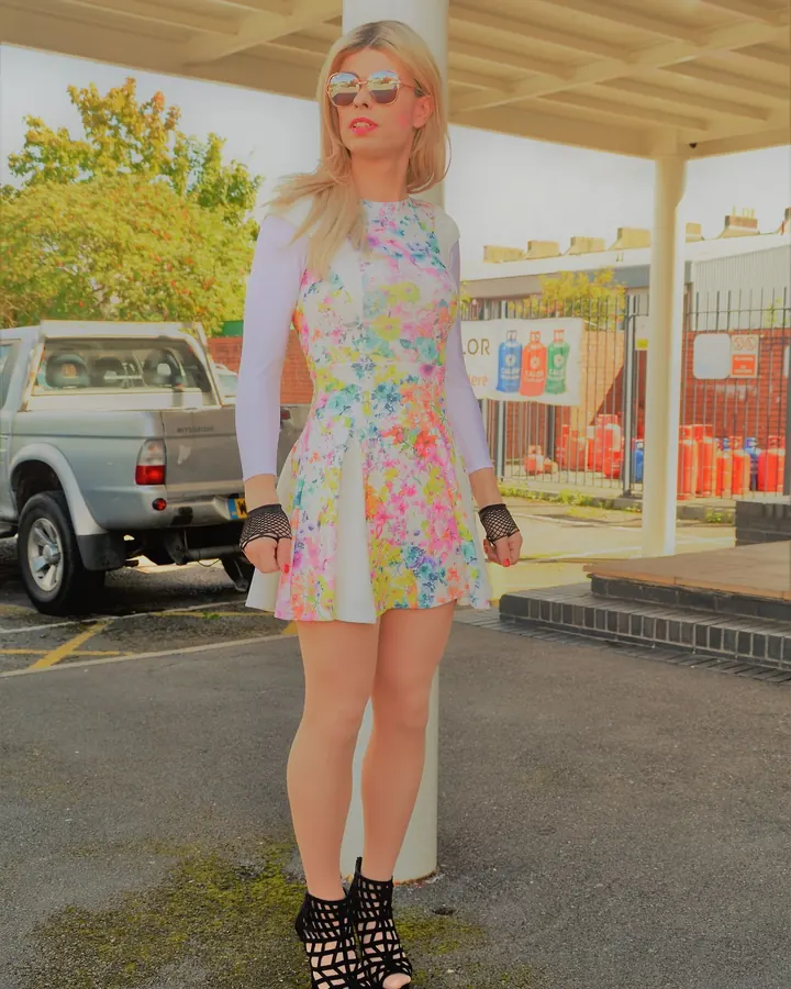 Femme blonde aux yeux marrons, en robe fleurie, posant devant un garage, lunettes de soleil et gants noirs.