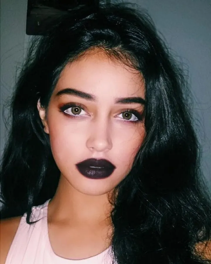 Femme aux yeux gris et cheveux bruns-noirs, en haut blanc, posant devant un mur vert.