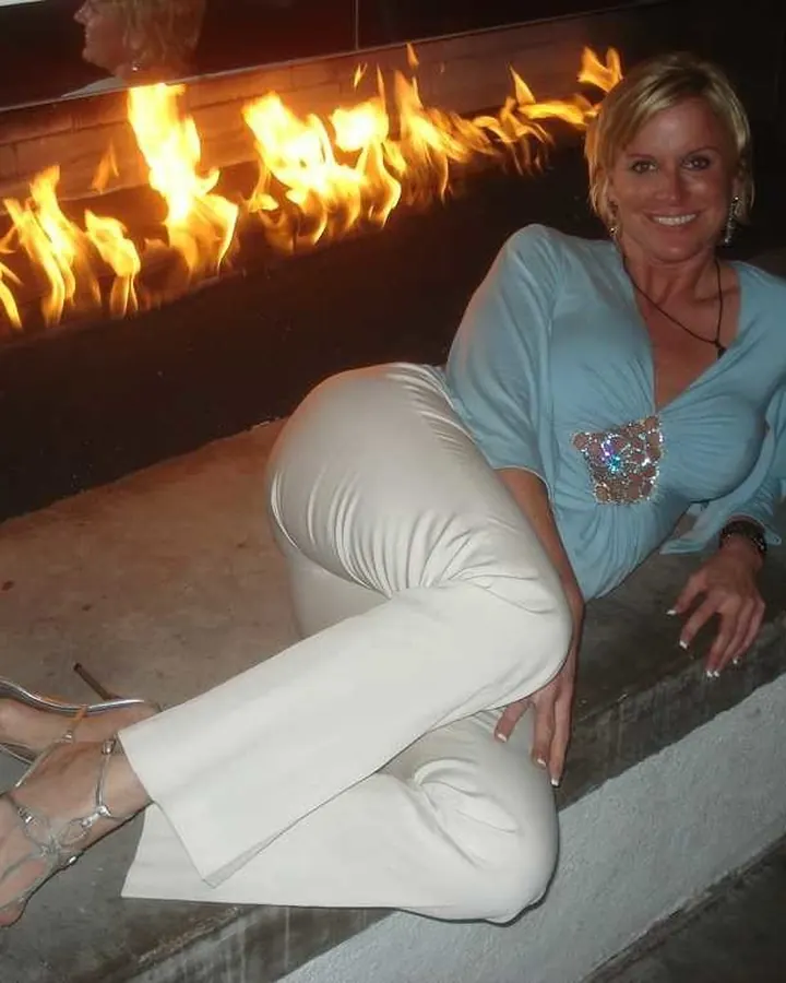 Femme blonde aux yeux bleus, en robe noire, posant sur un canapé, en collants résille et talons hauts.