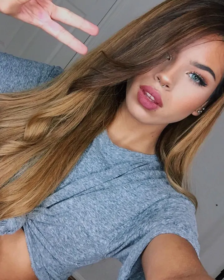 Jeune femme aux yeux bleus et cheveux blonds, en t-shirt gris, faisant un signe de la main tout en regardant l'appareil.