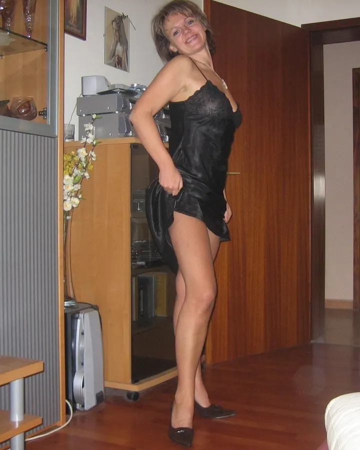 Femme aux yeux gris et cheveux blonds, en lingerie noire, souriant à l'appareil dans un salon.
