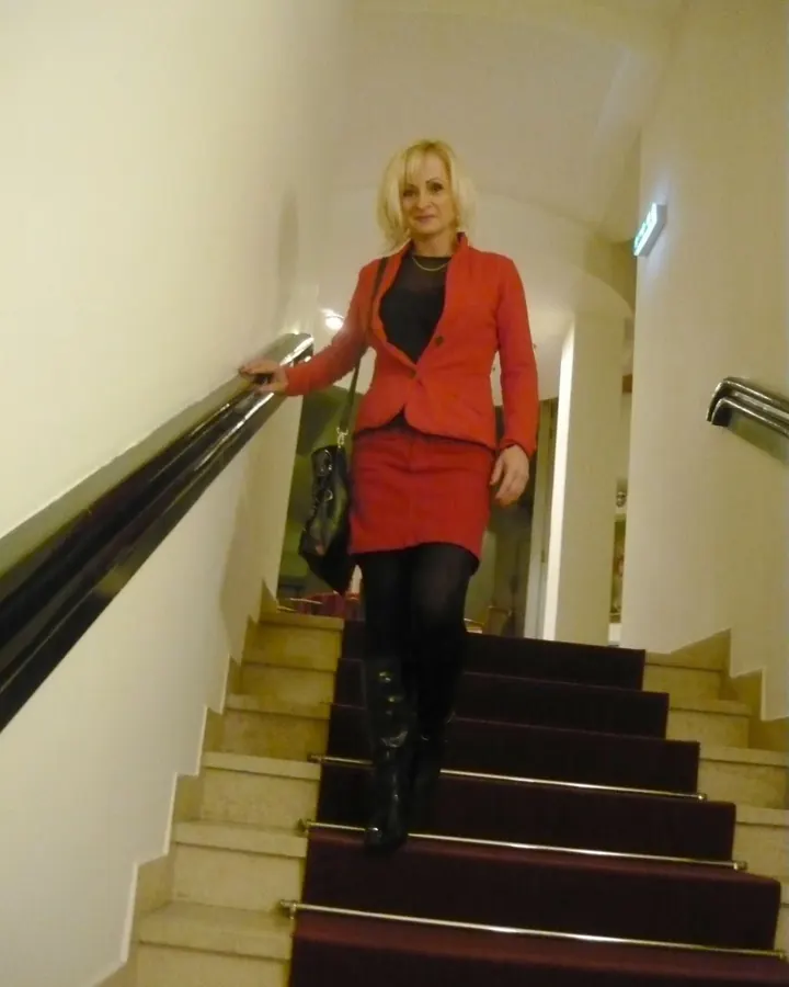 Femme blonde, souriante, descendant un escalier dans un hall, vêtue d'un tailleur rouge, collants noirs et bottes.