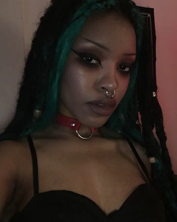 Femme aux cheveux noirs, en tenue sombre avec collier rouge, posant en intérieur, regard intense.