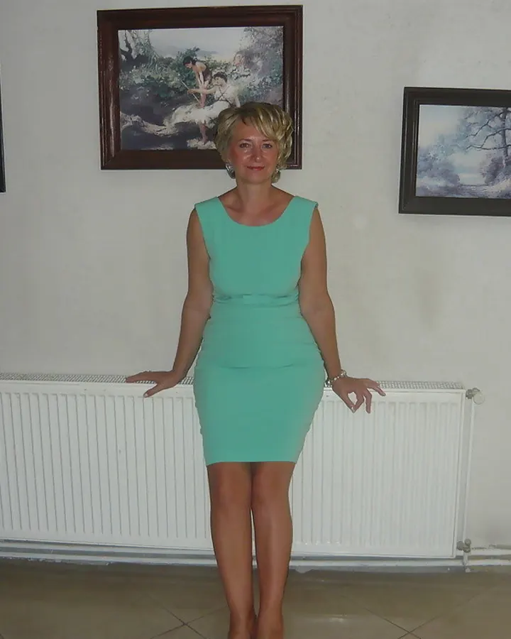 Femme aux cheveux blonds, en robe verte, posant dans un bureau en désordre, souriante.