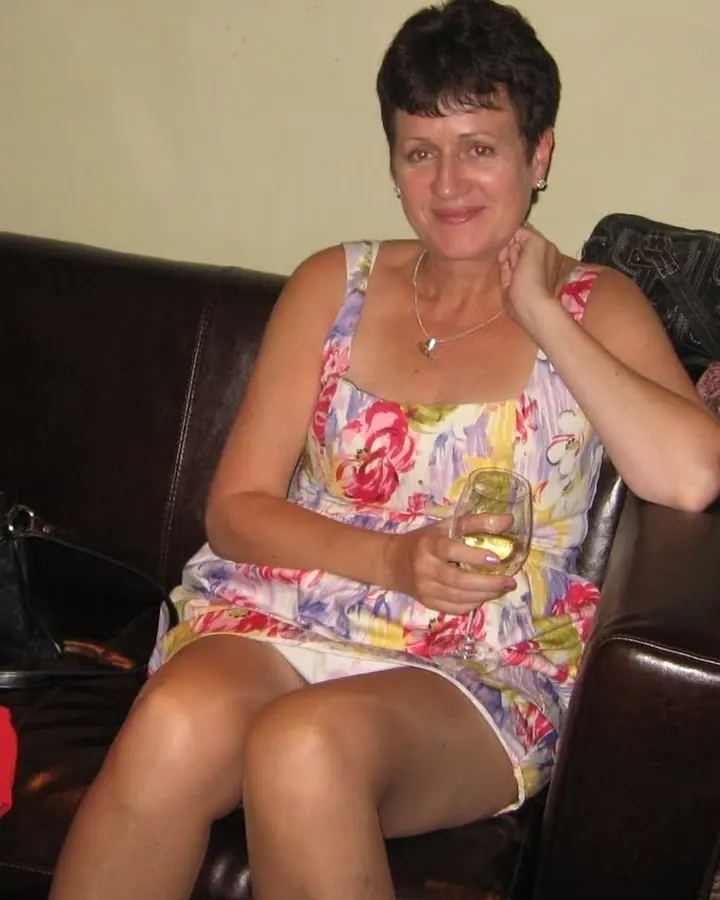 Femme aux cheveux bruns courts, portant une robe fleurie, assise sur un canapé en cuir, tenant un verre de vin blanc.